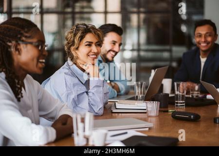 Lächelnde Geschäftsfrau schaut weg, während sie unter Kollegen am Konferenztisch sitzt Stockfoto