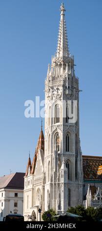 Der Turm der Mathias-Kirche in Buda: Der aus weißem Stein erbaute Turm leuchtet vor einem blauen Himmel, darunter die verzierten Dachziegel des Hauptgebäudes. Die Kirche der Himmelfahrt der Budaer Burg Stockfoto