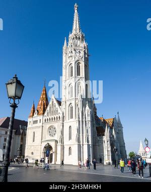 Die Fassade der Kirche Mariä Himmelfahrt der Budaer Burg oder der Matthias-Kirche: Der aus weißem Stein gebaute Turm leuchtet vor einem dunkelblauen Himmel die verzierten Dachziegel des Hauptgebäudes sind darunter. Stockfoto