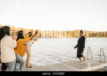 Frau tanzt auf dem Steg, während Freundinnen am Seeufer gegen den klaren Himmel jubeln Stockfoto