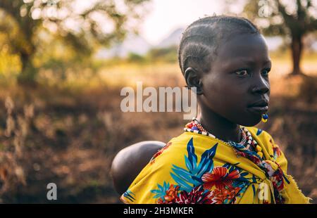 BOYA STAMM, SÜDSUDAN - 10. MÄRZ 2020: Mädchen in traditionellen bunten Gewand von Boya Tribe trägt Baby auf dem Rücken auf verschwommenem BA Stockfoto