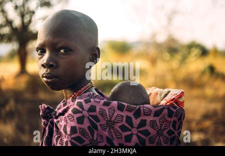 BOYA STAMM, SÜDSUDAN - 10. MÄRZ 2020: Mädchen in traditionellen bunten Gewand von Boya Tribe trägt Baby auf dem Rücken auf verschwommenem BA Stockfoto