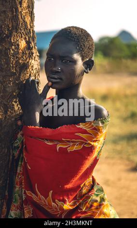 BOYA-STAMM, SÜDSUDAN - 10. MÄRZ 2020: Junge Frau in farbenfrohem Gewand, die Lippe berührt und wegschaut, während sie sich an Savanne lehnt Stockfoto