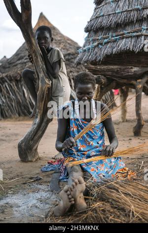 TOPOSA STAMM, SÜDSUDAN - 12. MÄRZ 2020: Frau sitzt auf schmutzigem Boden in der Nähe von Hütte und Kind und webt Produkt aus Stroh in vil Stockfoto