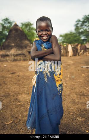 TOPOSA STAMM, SÜDSUDAN - MÄRZ 12, 2020: Glückliches Kind in bunten Tuch verpackt lächelnd für Kamera und Kreuzung Arme während livin Stockfoto