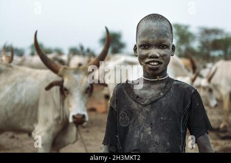 MUNDARI STAMM, SÜDSUDAN - 11. MÄRZ 2020: Junge Schäferhund in Lumpen lächeln vor der Kamera mit Interesse, während sie gegen Herde von stehen Stockfoto