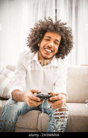 Netter Gamer spielt Spielkonsole, während er auf der Couch sitzt. Charmanter shaggy Araber in zerrissenen Jeans und einem weißen Hemd toothy Lächeln genießen Stockfoto