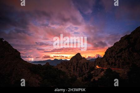 Sonnenaufgang über den Calanches von Piana auf Korsika mit Blitz in der Ferne und Autoampeln, die entlang der D81 fahren Stockfoto