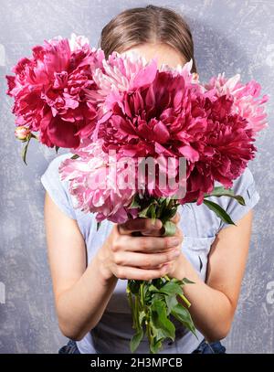 Frische, leuchtend rosa Pfingstrosen auf den Händen einer gesichtslosen jungen Frau in grauem T-Shirt auf grauer Nahaufnahme. Bild im trendigen Stil ohne Gesicht. Kreatives Bild für Mo Stockfoto