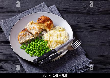 Gebratener Schweinelende und Sauerkraut auf einem Teller mit grünen Erbsen und Senf auf dunklem Holzboden, Draufsicht, Nahaufnahme Stockfoto