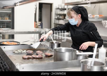 Frau Chefin in schützender Gesichtsmaske bereitet Essen in der Küche eines Restaurants oder Hotels zu. Coronavirus-Präventionskonzept. Stockfoto