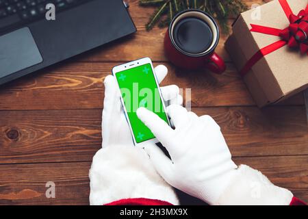 Der weihnachtsmann hat mit Handschuhen ein Telefon in der Hand. Chromakey. Daneben befindet sich ein Laptop, eine Tasse, ein Geschenk und Tannenzweige. Draufsicht. Stockfoto