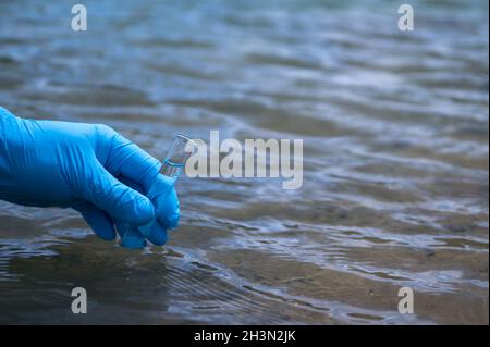 Die Hand eines Mannes in einem blauen Handschuh nimmt eine Nahaufnahme des Wassers in ein Reagenzglas, um die Wasserverschmutzung zu messen. Hintergrund. Stockfoto