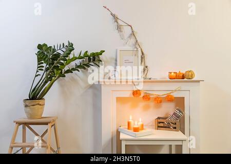 Rahmen mit Schild 'Happy Halloween'. Zamioculcas Pflanzen im Tontopf auf dem Stuhl. Girlande von Skeletten hängt an der Wand. Girlande aus Kunststoff-Kürbissen hängt von Stockfoto