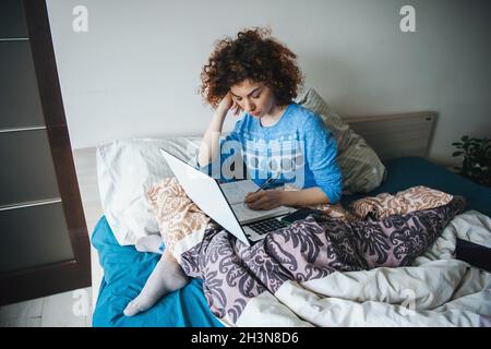 Lockige Frau, die aus der Ferne studiert und in einem unordentlichen Bett sitzt. Moderner Frauenleben. Stockfoto