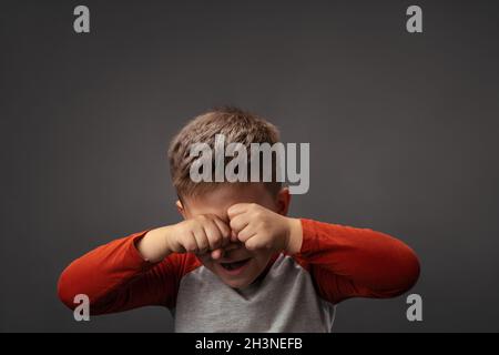 Weinender kleiner Junge drückt traurige Emotionen aus. Verärgert Kind reibt Augen mit Fäusten isoliert auf grauem Hintergrund. Problemkonzept Stockfoto