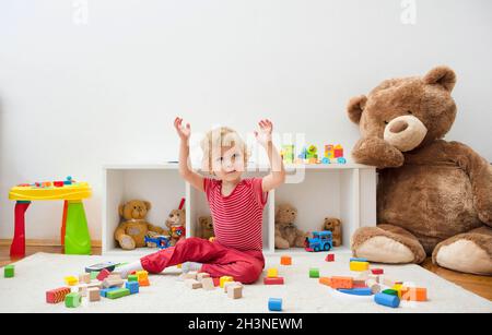 Süßer, glücklicher Junge, der zu Hause mit seinem riesigen Teddybären und bunten Holzblöcken und Spielzeug auf dem Boden Spaß hat. Stockfoto