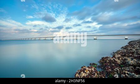 Lange Brücke über glasig glattes Wasser geht bis in die Unendlichkeit über den herrlichen Ozean unter dem wunderschönen bunten Himmel in Zeeland, den Niederlanden Stockfoto