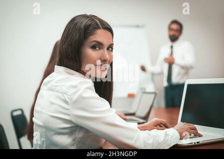 Die hübsche Geschäftsfrau blickt auf die Kamera zurück, die am Arbeitsplatz am Laptop arbeitet. Männlicher Sprecher, der die Präsentation des Projektstandens hält Stockfoto