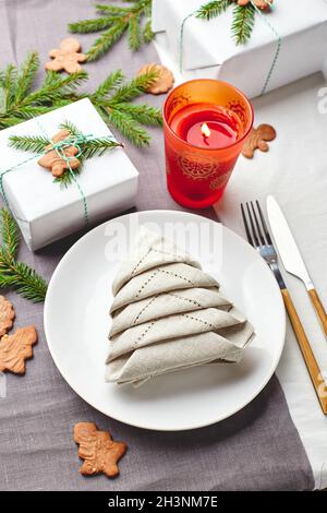 Serviette in Form eines Weihnachtsbaums auf einem Teller auf weißer Tischdecke mit Geschenken und Dekorationen mit Tannenzweigen und Lebkuchen Stockfoto