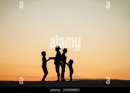 Silhouette einer Familie bei Sonnenuntergang am Strand. Mama und Papa stehen mit dem Rücken zueinander und halten die Hände mit ihren Söhnen. Stockfoto