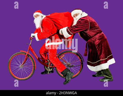 Zwei fröhliche Weihnachtsmänner in Neujahrsmützen und roten Anzügen fahren mit einer Geschenktüte auf dem Fahrrad. Isolieren. Frohes Neues Jahr 2022. Frohe Weihnachten. Konz Stockfoto