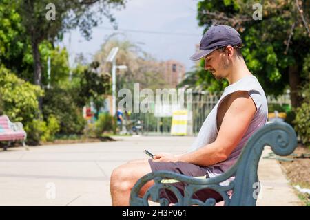 Junger Mann mit Mütze, der am sonnigen Tag auf der Parkbank auf das Mobiltelefon schaut. Millenial Junge auf ärmellosem Hemd und Shorts mit Smartphone im Freien Stockfoto