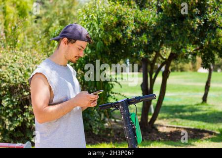 Junger Mann mit Smartphone per Roller im Park am sonnigen Tag. Alternativer Pendelverkehr, mobile App-Konzepte Stockfoto