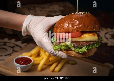 Hand in einem Einweg-Glow mit frischem Craft Beef Burger mit gebratenem Käse, serviert mit frischem Salat, Pommes frites und Tomaten Stockfoto