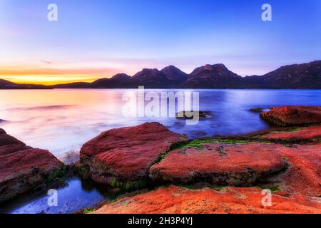 Malerischer Sonnenaufgang über der Freycinet Peninsula-Bergkette durch die Coles Bay mit roten Flechten bedeckte Granitfelsen. Stockfoto