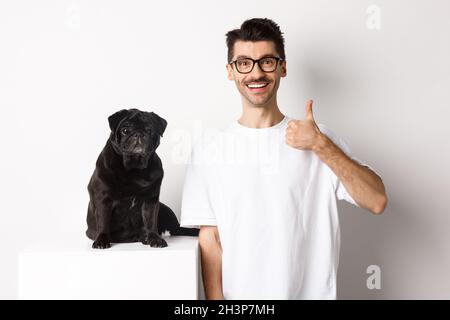 Bild von Hundebesitzer und niedlichem schwarzen Mops, der die Kamera anschaut, Mann, der mit Daumen nach oben zeigt, etwas empfiehlt, überstehend Stockfoto