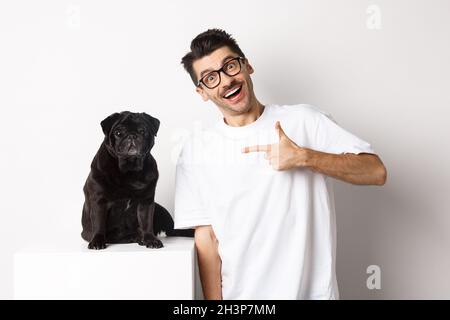 Glücklicher junger Mann, der seinen niedlichen Hund zeigt, mit dem Finger auf schwarzen Mops zeigt und lächelt und auf weißem Hintergrund steht Stockfoto