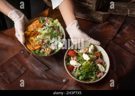 Zwei Hände mit Schüsseln Gemüsesalat mit Lachs, Mozzarella-Käse und gegrillten Scheiben von Früchten steht auf einem Tisch bedeckt wi Stockfoto