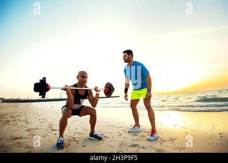 Der gutaussehende Glatze hockt mit einer Langhantel am Strand mit seinem Freund. Stockfoto