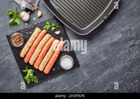 Rohe Fleischwürste auf einem Schieferbrett, mit Kräutern und Gewürzen und einer quadratischen Grillpfanne. Blick von oben Stockfoto