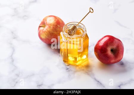 Glas mit flüssigem Honig aus Blumenmuster mit Honiglöffel aus Metall und roten Äpfeln auf hellem Marmorhintergrund Stockfoto