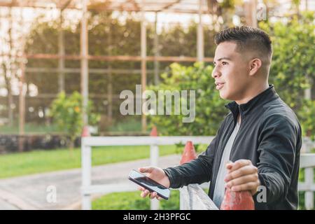 Unternehmungslustiger junger Mann neben dem weißen Holzzaun mit seinem Handy in den Händen, der geradeaus blickt, glücklich über seine Leistungen nachdenkt, Stockfoto