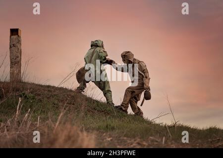 Ein Mann in einer Gasmaske und einem chemischen Schutzanzug hilft einer anderen Person zu gehen. Post-Apokalypse nach Atomkrieg und Strahlung Stockfoto