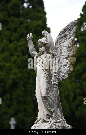 Eine Steinskulptur eines geflügelten Engels auf einem Friedhof, der ein Horn in der linken Hand hält, mit Bäumen und einem Kreuz im Hintergrund Stockfoto