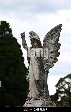 Steinskulptur eines Engels, der ein Horn auf einem Friedhof hält, mit einem hellen, aber bewölkten Himmel im Hintergrund Stockfoto