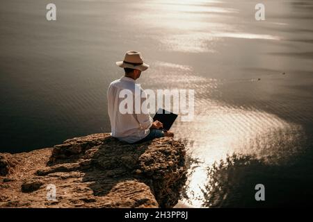 Digitaler Nomade, Mann im Hut, ein Geschäftsmann mit einem Laptop sitzt auf den Felsen am Meer während des Sonnenuntergangs, macht eine Geschäftstransaktion online von einem Stockfoto