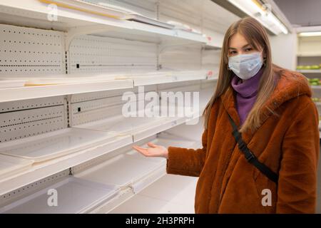 Junge Frau in medizinischer Gesichtsmaske und leeren Regalen im Supermarkt. Stockfoto