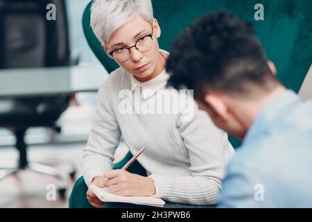 Frau mit Stift und Notizbuch in der Hand schreibt und Mann sitzt zusammen auf einem Stuhl. Umfrage zu Journalismus, Interview und Personalbeschaffung Co Stockfoto