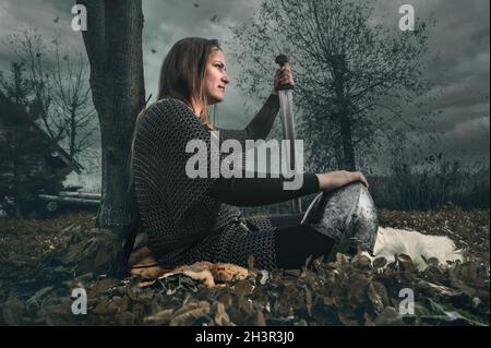 Portrait der Frau in Bild des mittelalterlichen Kriegers unter dramatischem Himmel. Schöne Frau in mittelalterlichen Kettenpost sitzt nach der Schlacht Stockfoto