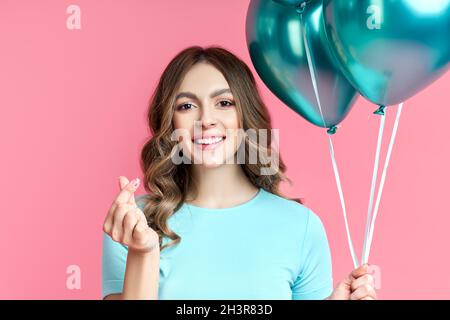 Ziemlich lächelnde Frau macht Mini Herz koreanischen Liebe Symbol durch Finger und hält blaue Ballons auf rosa Hintergrund Stockfoto