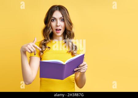 Porträt der erstaunt überrascht Frau zeigt auf Buch mit Kopieplatz auf gelbem Hintergrund Stockfoto