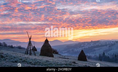 Der erste Herbstfrost auf der Weide mit Heuhaufen und majestätischem Sonnenaufgang am Rande des Bergdorfes. Stockfoto