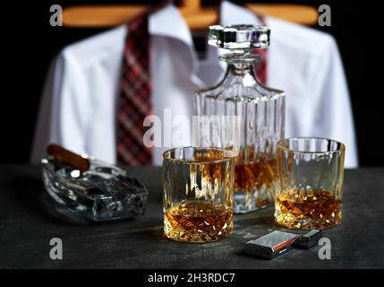 Stillleben mit Flasche und zwei Gläsern Whisky auf einer Steinplatte in einer Bar. Zigarre mit weißem Hemd und roter Krawatte auf schwarzem Bac Stockfoto