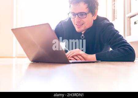 Auf dem Holzboden sitzt ein fröhlicher junger Mann mit einem modernen Laptop Stockfoto