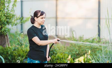 Frau, die den Garten aus Schlauch wässert. Weibchen sprühen Wasser auf Gemüse mit einem Gartenschlauch. Eine glückliche Frau mit einem Schlauch kümmert sich um den Garten. Stockfoto